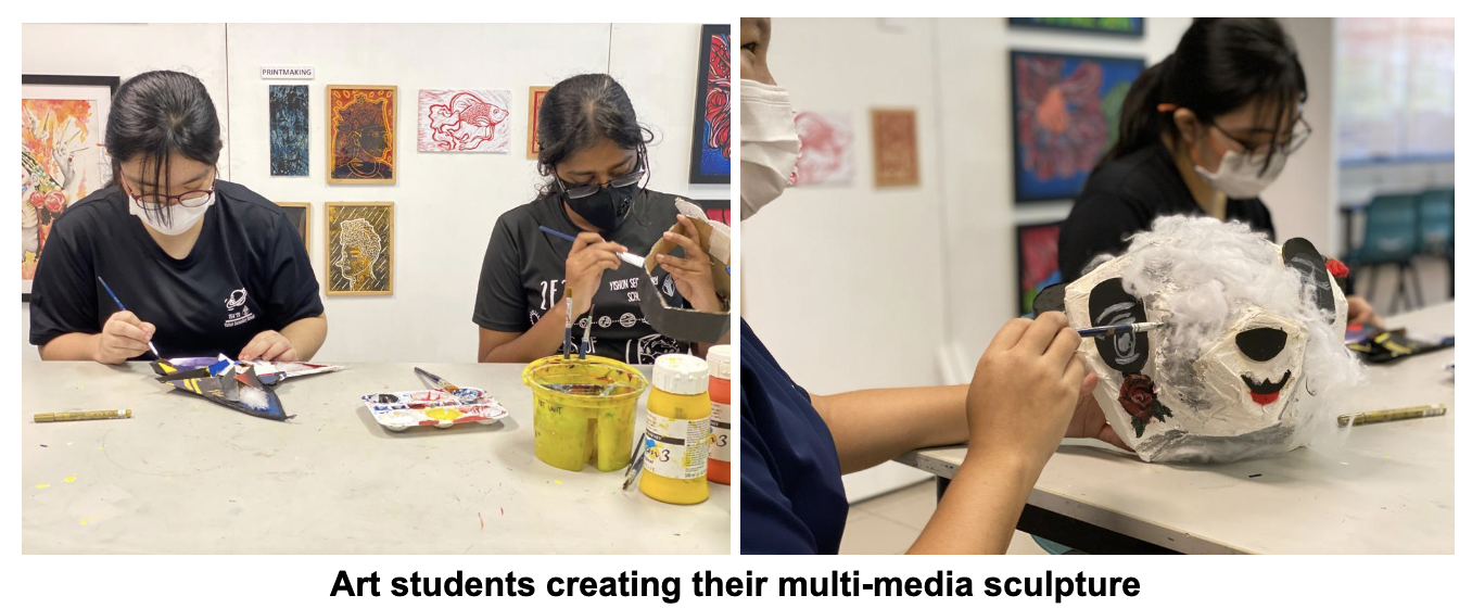Two masked students paint at a table with artwork on walls; another paints a white panda-like paper mache mask.